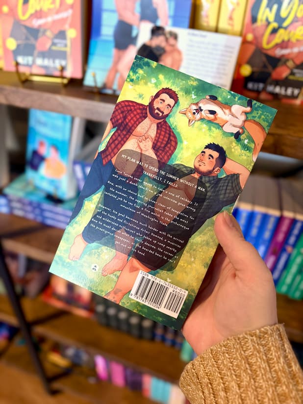 A hand holding a copy of 'Cubs & Campfires' by Dylan Drakes, featuring an illustration of two men with their shirts undone and a dog lying down in the grass.