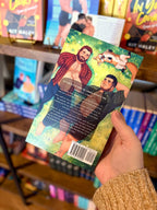 A hand holding a copy of 'Cubs & Campfires' by Dylan Drakes, featuring an illustration of two men with their shirts undone and a dog lying down in the grass.