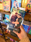 A hand holding a copy of 'Cubs & Campfires' by Dylan Drakes, featuring and illustration of two men hugging in a forest setting.