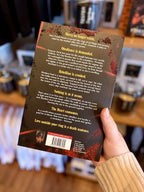 A hand holding the back of a paperback copy of 'Daggermouth' by H. M, featuring a black and red background with white and yellow text.
