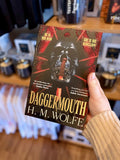 A hand holding a paperback copy of 'Daggermouth' by H. M, featuring a black and red background with a black skull in the centre.