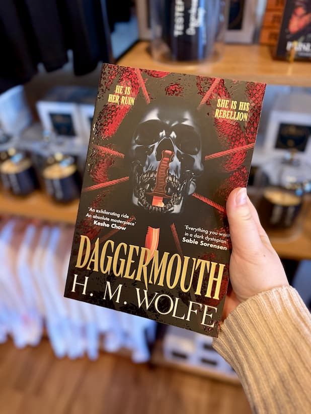 A hand holding a paperback copy of 'Daggermouth' by H. M, featuring a black and red background with a black skull in the centre.