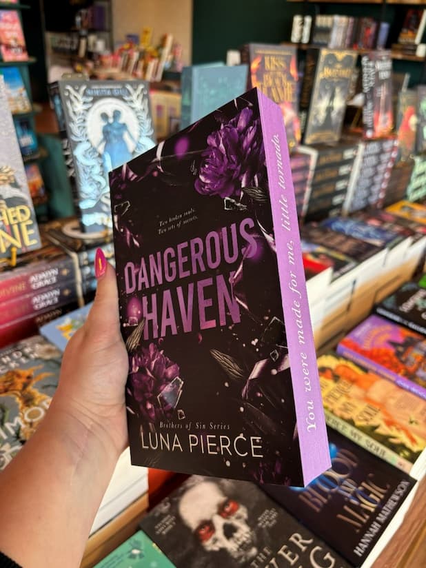 A hand with pink nail polish holding a copy of 'Dangerous Haven' by Luna Pierce, featuring a dark gray cover with purple flowers and purple title text. The side shows purple sprayed edges with the quote 'You were made for me, little tornado.'