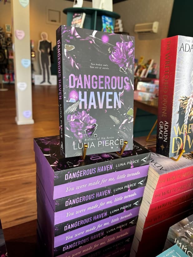 A stack of copies of the book 'Dangerous Haven' by Luna Pierce, with one book standing upright on top of the others in a bookstore display. The cover is dark gray with purple flowers and purple title text, and the stacked copies show purple sprayed edges with the quote 'You were made for me, little tornado.'
