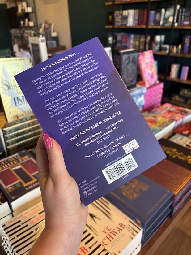A hand with pink nail polish holding the back of a copy of 'Dangerously Ours' by Krista and Becca Ritchie with a purple background and white text.