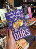 A hand with pink nail polish holding a copy of 'Dangerously Ours' by Krista and Becca Ritchie with a gold heart locket open with golden spider's web protruding from inside. The locket is twisted and purple flowers and thorns on a purple background.