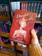 A hand holding a paperback copy of 'Daughter of No Worlds', featuring a woman of stone holding a heart and gold elements on a red background.