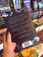 A hand with dark glittery nail polish holds the back cover of a book titled 'Daybreak' by Autumn Woods in a bookstore, with other books visible on the shelves in the background.
