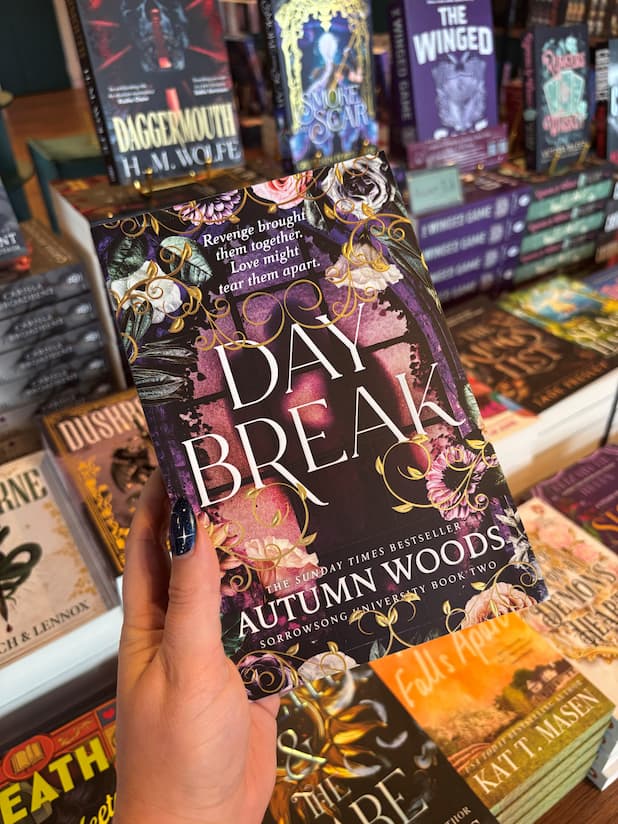 A hand holds a copy of the book 'Daybreak' by Autumn Woods in a bookstore, with other books visible on the shelves in the background.