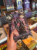 A hand holds a copy of the book 'Daybreak' by Autumn Woods in a bookstore, with other books visible on the shelves in the background.