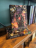 The book 'Daybreak' by Autumn Woods displayed on a wooden bookshelf next to other books, with a dark teal wall in the background.
