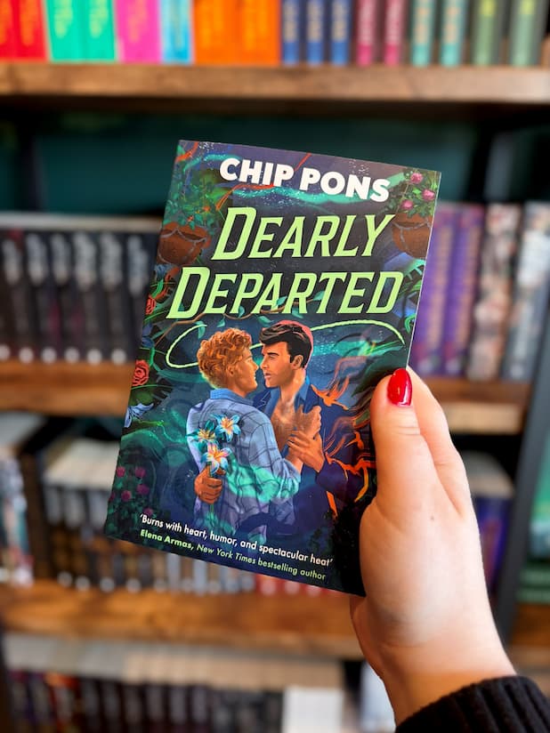 A hand with red nail polish holds a paperback book titled 'Dearly Departed' by Chip Pons. A sexy Hades-inspired gay rom-com. The book cover features an illustration of a male couple embracing with flowers decorating around them in a variety of colours.