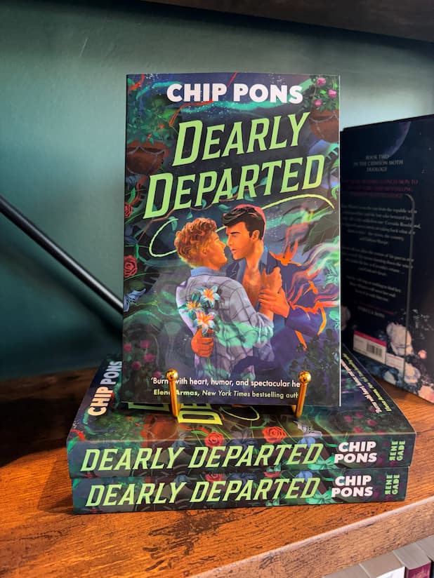 A copy of the book 'Dearly Departed' by Chip Pons displayed on a wooden bookshelf, standing upright on a small gold book stand next to other copies of the book.