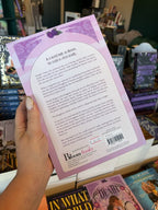 A hand with nude holding the back of a copy of 'Death' by Kristin Cast, featuring a pastel purple background surrounded by floral elements and purple text.
