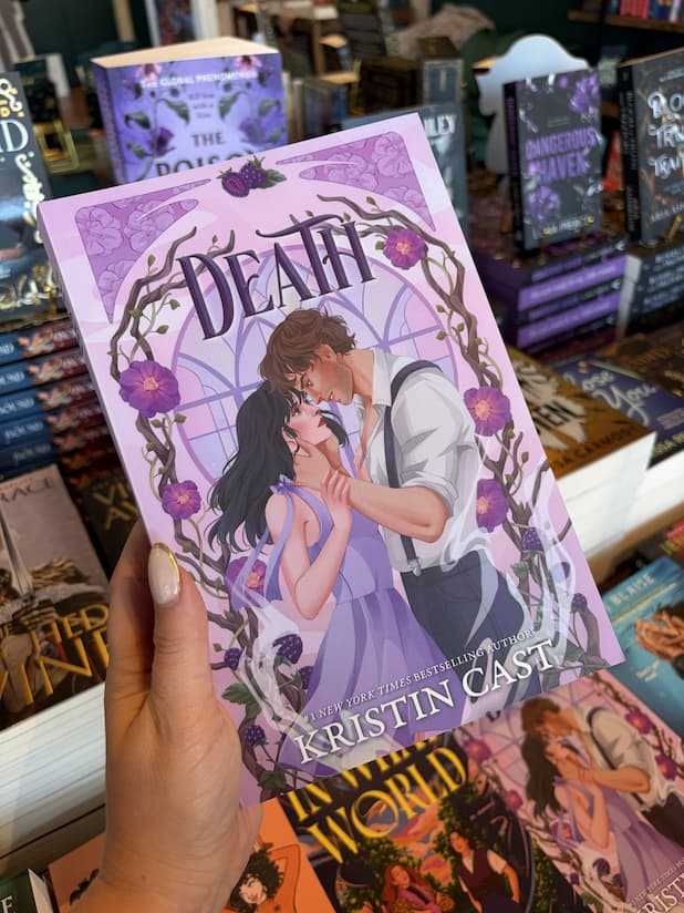 A hand with nude holding a copy of 'Death' by Kristin Cast, featuring a romantic illustration of two characters looking deeply into each other's eyes, he is cradling her face between his hands and they are surrounded by floral elements.