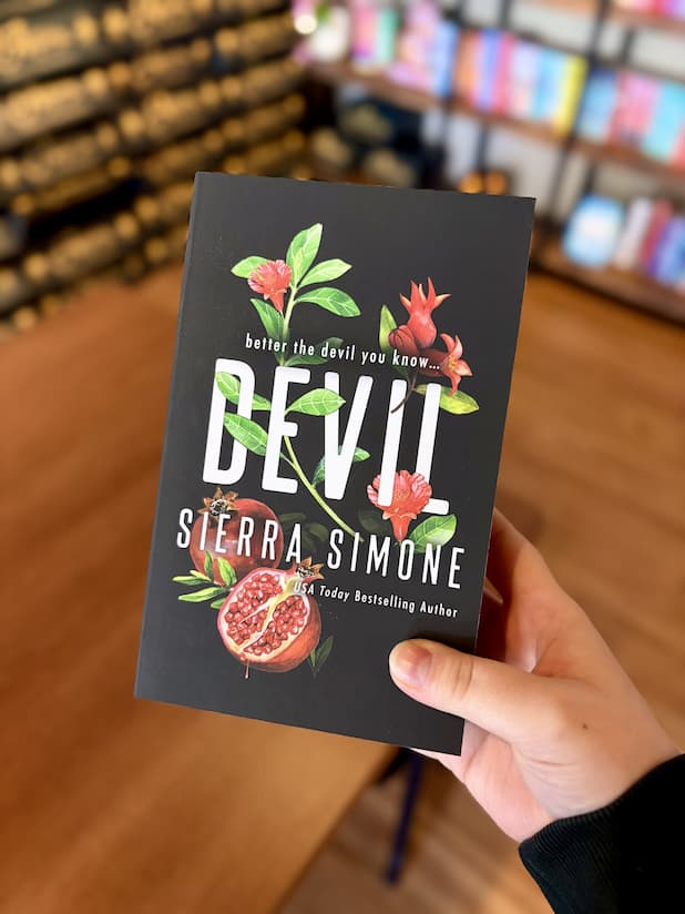 A hand holding a paperback copy of 'Devil' by Sierra Simone, featuring a black background with red flowers and pomegranates illustrated between the title font.