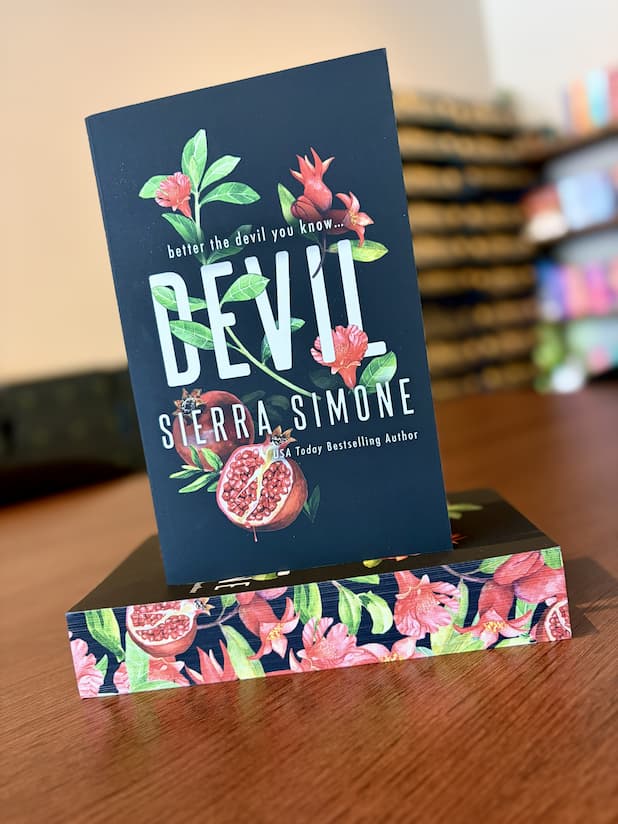 A small stack of 'Devil' by Sierra Simone, featuring a black background with red flowers and pomegranates illustrated between the title font. The lying down book shows the sprayed edges that contain the same flower and pomegranate illustrations.