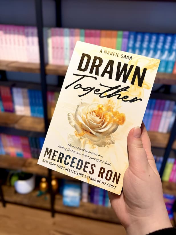 A hand holding a copy of 'Drawn Together' by Mercedes Ron with a rose design and golden accents on a white background.
