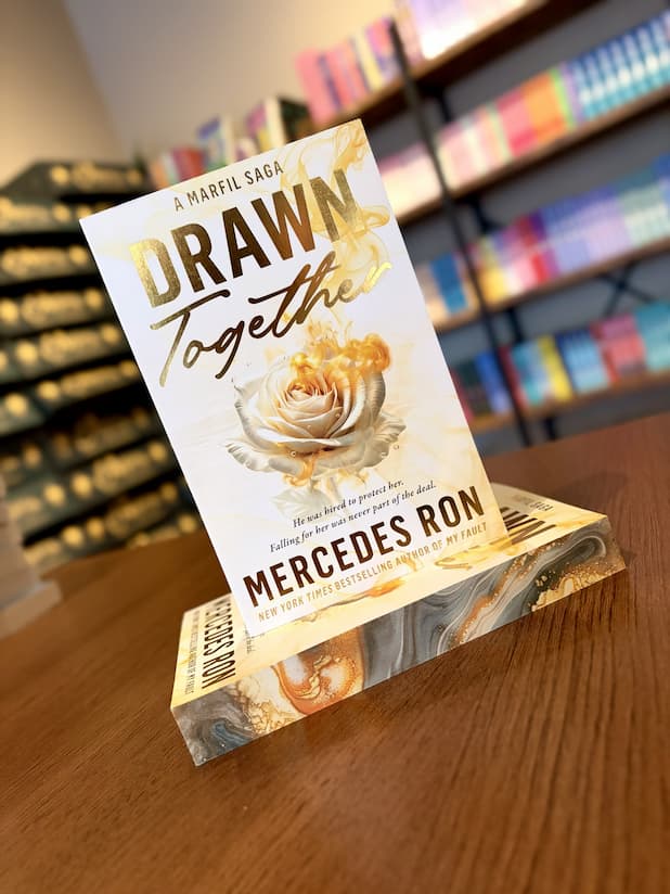 A small stack of 'Drawn Together' by Mercedes Ron with a rose design and golden accents on a white background with marbled sprayed edges.
