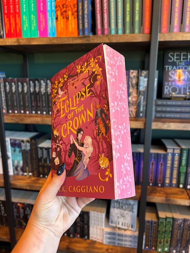 A hand with blue starry nail polish holds the Special Edition Book of 'Eclipse of the Crown' by A.K. Caggiano with a purple background and characters embracing in a dance on the cover with pink floral edges in front of a blurry bookshelf