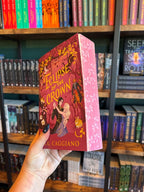 A hand with blue starry nail polish holds the Special Edition Book of 'Eclipse of the Crown' by A.K. Caggiano with a purple background and characters embracing in a dance on the cover with pink floral edges in front of a blurry bookshelf