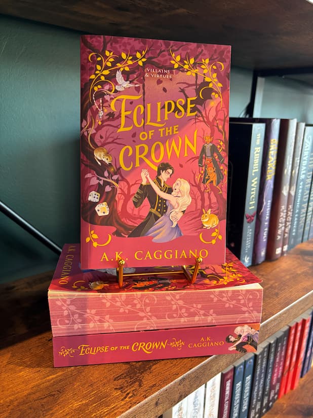 A copy of the book 'Eclipse of the Crown' by A.K. Caggiano displayed on a wooden bookshelf, standing upright on a small gold book stand next to other copies of the book.