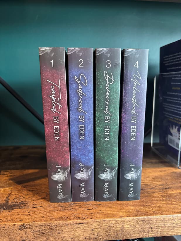 Four paperback copies of the book series 'Eden' by Jade May, showing the spines of 'Tempted by Eden', 'Seduced by Eden', 'Devoured by Eden' and 'Unleashed by Eden' lined up on a wooden shelf against a green wall.