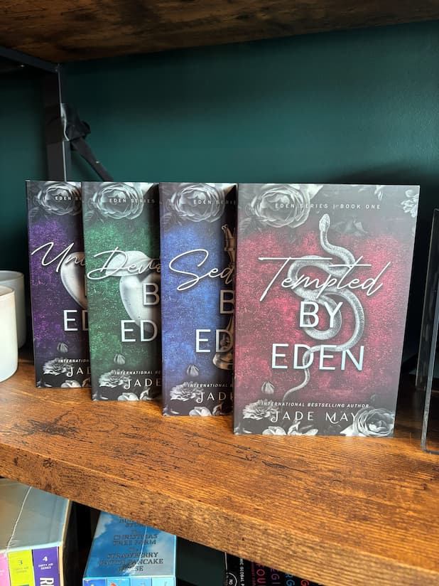 Four paperback copies of the book series 'Eden' by Jade May, showing the covers of 'Tempted by Eden', 'Seduced by Eden', 'Devoured by Eden' and 'Unleashed by Eden' lined up on a wooden shelf against a green wall.