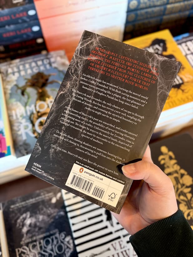 A hand holding the back of a copy of 'Eldritch' by Keri Lake, featuring a black and grey forest with white and red text.