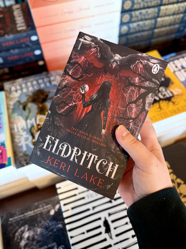 A hand holding a copy of 'Eldritch' by Keri Lake with a woman in a dark dress walking in a forrest carrying a skull and a large black bird flies toward her.