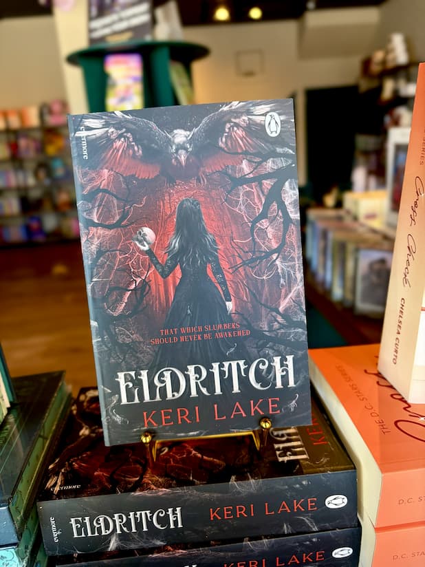 A stack of 'Eldritch' by Keri Lake with a woman in a dark dress walking in a forrest carrying a skull and a large black bird flies toward her.
