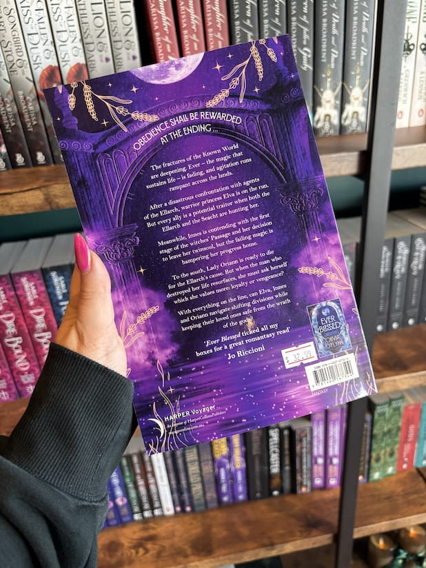 A hand with pink nail polish holding the back of a copy of 'Ever Cursed' by Olivia O'Fynn, featuring a purple celestial archway with a full moon, gold decorative flourishes, and white text.
