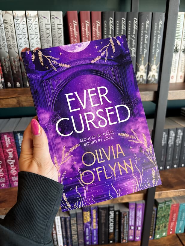 A hand with pink nail polish holding a copy of 'Ever Cursed' by Olivia O'Fynn, featuring a purple celestial archway with a full moon, gold decorative flourishes, and white and gold title text.