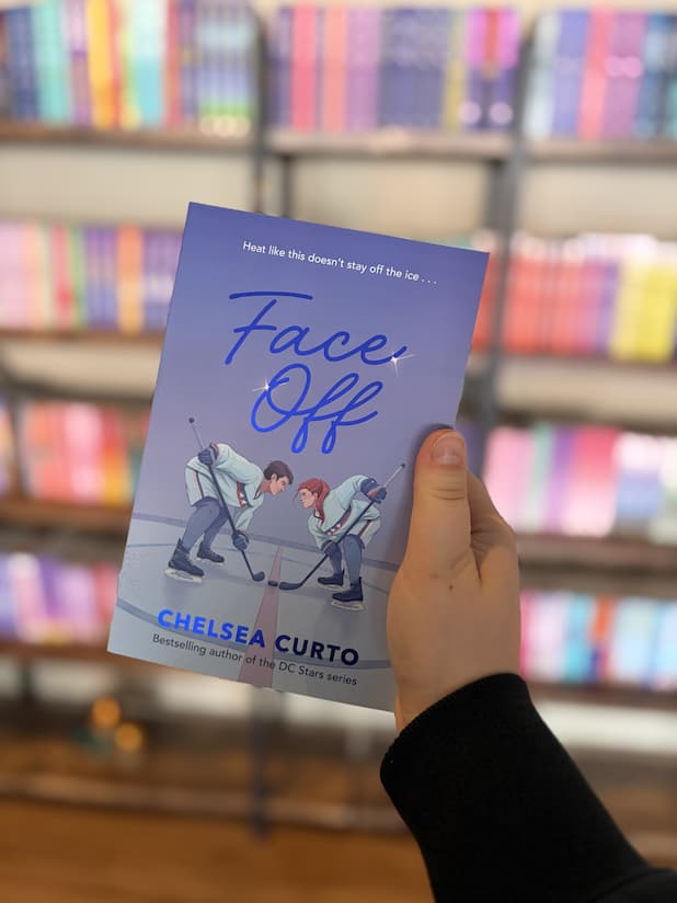 A hand holds a copy of 'Face Off' from The D.C. Stars Series by Chelsea Curto, featuring two hockey players on an ice rink.