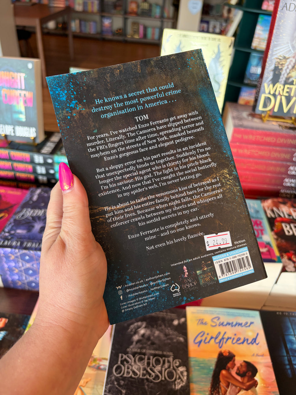 A hand with pink nail polish holds the back cover of the book titled 'Fallen Rook' by L.J. Shen in a bookshop surrounded by other blurry books. The cover features a dark background with blue paint spashes in the corner of the book and white text for the blurb.