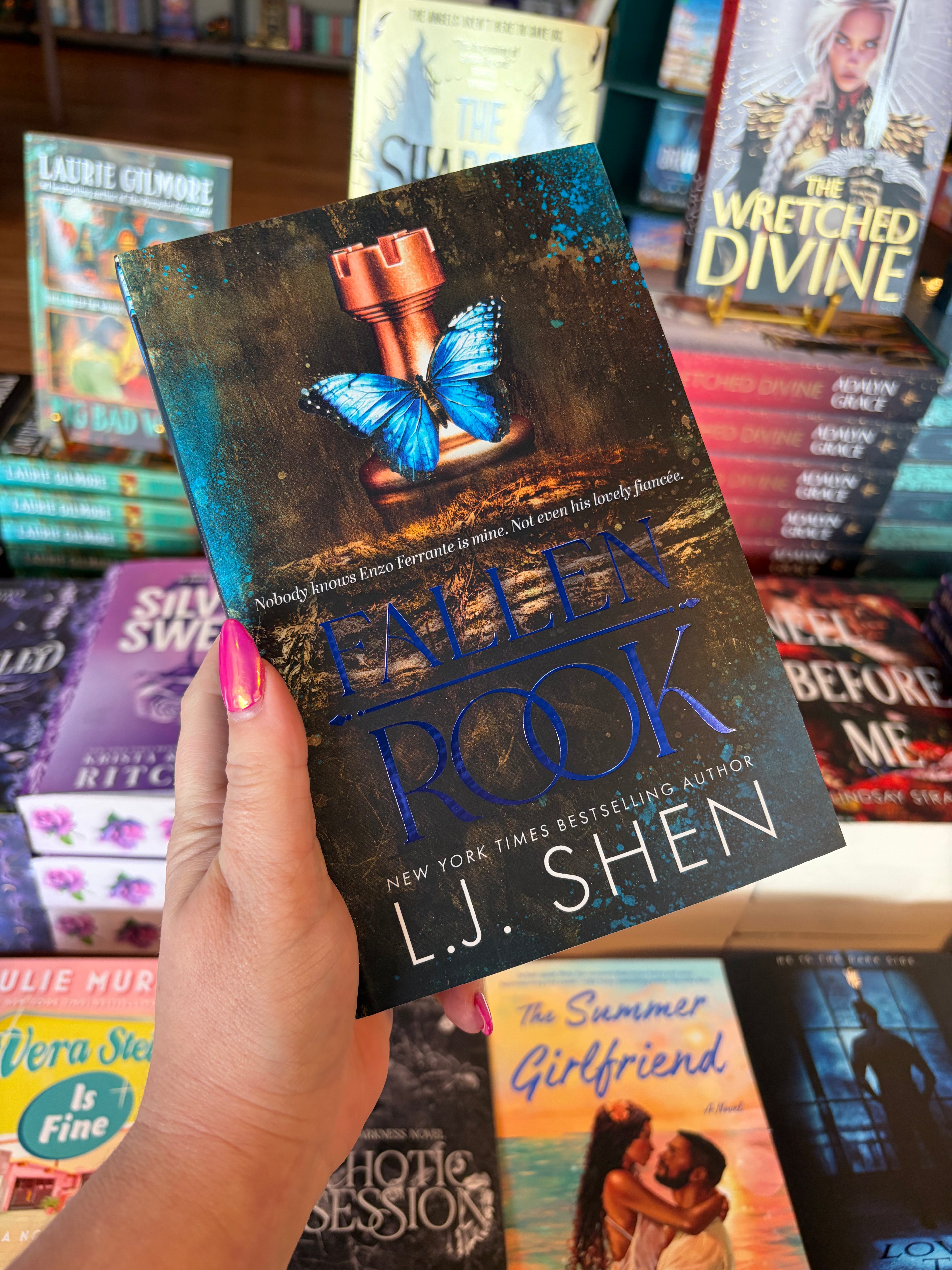 A hand with pink nail polish holds a book titled 'Fallen Rook' by L.J. Shen in a bookshop surrounded by other blurry books. The cover features a blue butterfly sitting on a copper rook chess piece on a log with moss and blue paint spashes in the corner of the book.