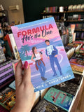 A hand with pink nail polish holding a copy of 'Formula: He's the One' featuring two characters on a race track with palm trees and cityscape in the background.