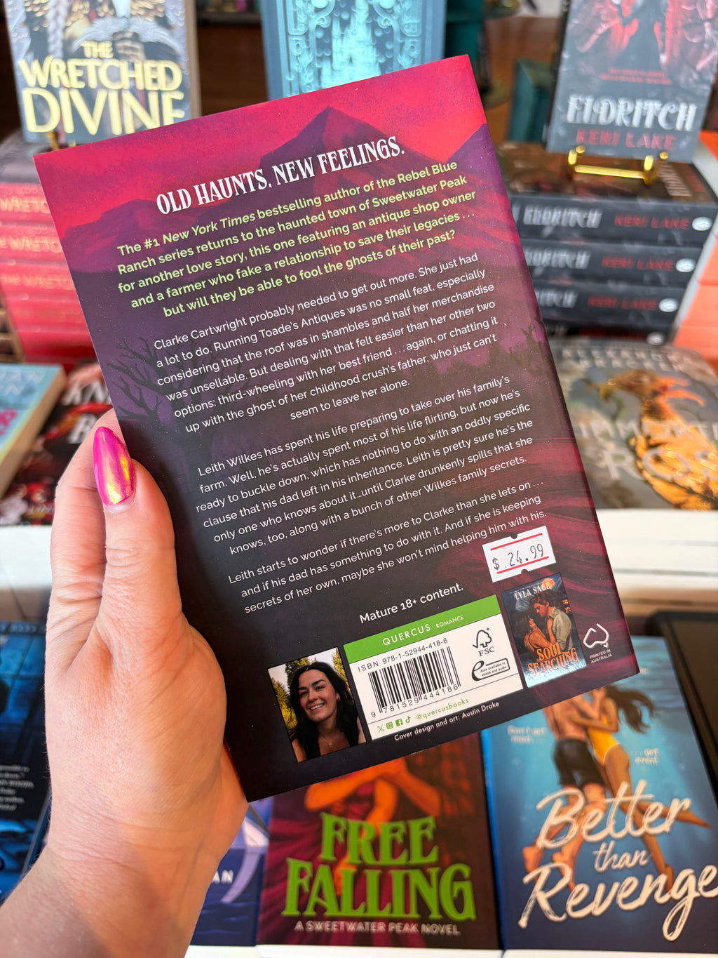 A hand with pink glitter nail polish holds up the back cover of the paperback book Free Falling by Lyla Sage. The book cover features a dark pink and purple background with white text for the blurb.