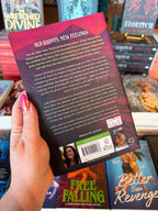 A hand with pink glitter nail polish holds up the back cover of the paperback book Free Falling by Lyla Sage. The book cover features a dark pink and purple background with white text for the blurb.