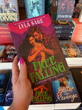 A hand with pink nail polish holds up a copy of the book 'Free Falling' by Lyla Sage in a bookstore setting, with other copies of the book and shelves visible in the background. The cover features a couple embracing her back to his chest, with his hand on her shoulder and waist. they are in front of a pink mountain landscape. 