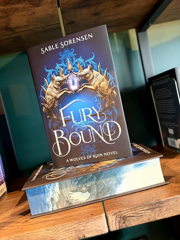 The book 'Fury Bound' by Sable Sorensen, a Wolves of Ruin novel, displayed on a wooden shelf. The cover features a dark blue background with a golden wolf and a glowing blue gem, and the title is written in large white letters.