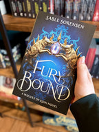 A hand holding a paperback copy of 'Fury Bound' by Sable Sorensen featuring two metal wolf heads holding a crystal against a blue background.