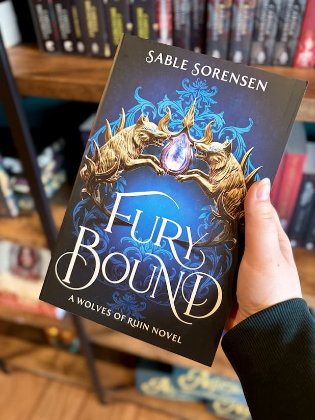 A hand holding a paperback copy of 'Fury Bound' by Sable Sorensen featuring two metal wolf heads holding a crystal against a blue background.