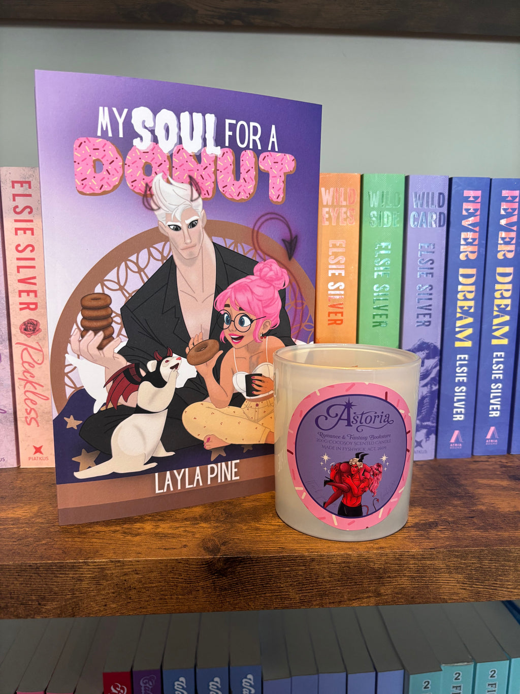 A candle in a frosted glass jar with a purple and pink label reading 'Astoria' sits on a wooden shelf next to a matching book cover titled 'My Soul for a Donut' by Layla Pine, with other books in the background.