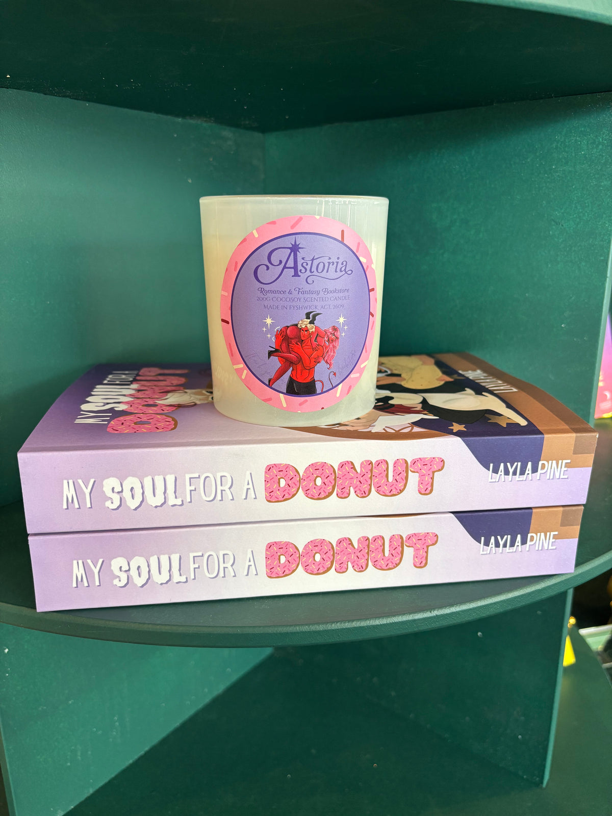 A frosted glass candle with a purple and pink circular label sits on top of two copies of the book 'My Soul for a Donut' by Layla Pine, displayed on a dark green shelf.