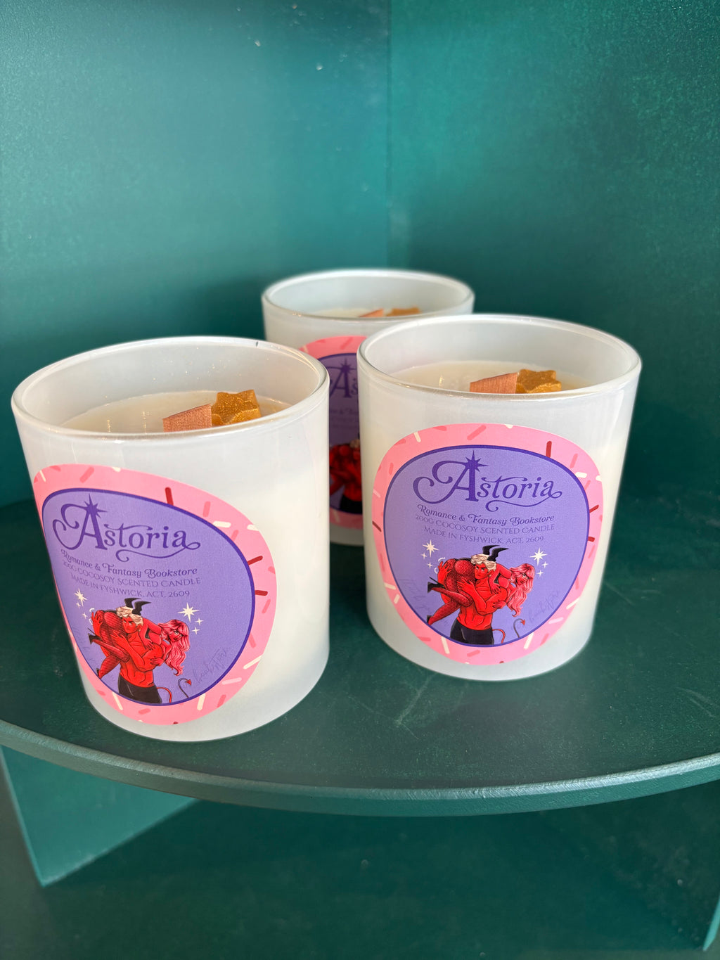 Three white soy candles in clear glass jars with purple and pink Astoria labels, displayed on a teal shelf against a teal background.