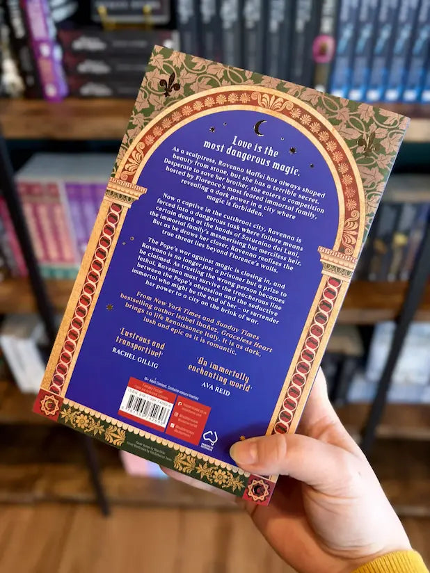 A hand holding the back of a paperback copy of the book 'Graceless Heart' by Isabel Ibánez, with a blue backgroun and gold ornate detailing.