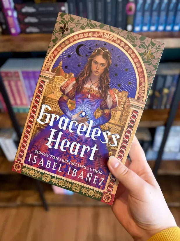 A hand holding a paperback copy of the book 'Graceless Heart' by Isabel Ibánez, with a woman in a historical costume and cityscape background.