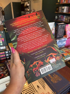 A hand with pink nail polish holding the back of a copy of 'Green & Deadly Things' by Jenn Lyons, featuring a red sun and tropical green leaves with white and yellow text.
