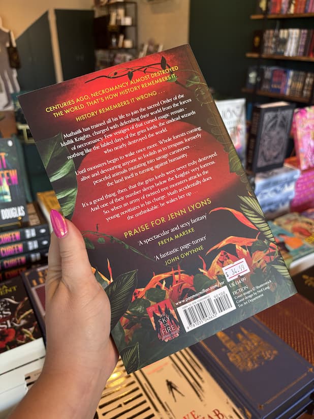 A hand with pink nail polish holding the back of a copy of 'Green & Deadly Things' by Jenn Lyons, featuring a red sun and tropical green leaves with white and yellow text.